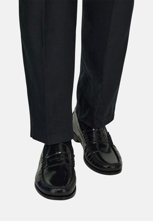 Mocassins en cuir noir brillant portés avec un pantalon de costume noir, présentant des vues de face et de profil sur un fond clair uni.