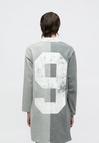 Pilka didelė laisvo stiliaus megztinis su ilgomis rankovėmis, ant nugaros yra didelis dėvėtas baltas skaičius 9. Dizainas suskirstytas į dvi pilkos spalvos atspalvių dalis.