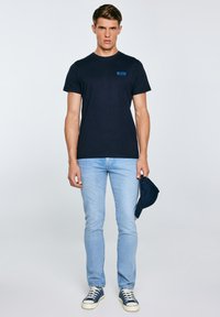 Mees, kellel on seljas must "BIG STAR" T-särk, helesinised teksad ja mereväesinised tossud, hoiab paremas käes sobivat mereväesinist nokamütsi.