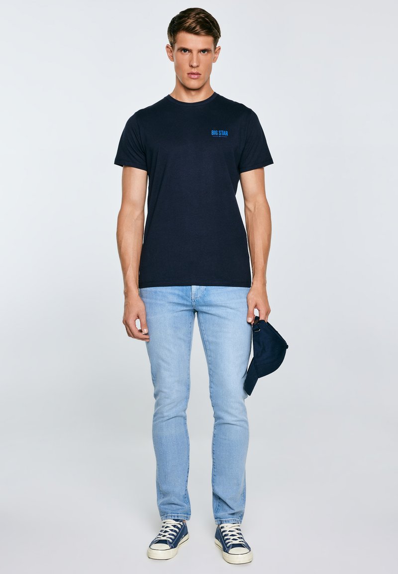 Mees, kellel on seljas must "BIG STAR" T-särk, helesinised teksad ja mereväesinised tossud, hoiab paremas käes sobivat mereväesinist nokamütsi.