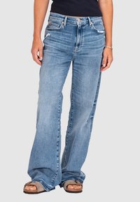 Jasnoniebieskie dżinsy o szerokich nogawkach z denimu o wyblakłym wykończeniu. Posiadają postrzępione detale i wysoki stan. Nosi się je z brązowymi sandałami.