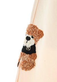 Parche de oso de peluche sobre una tela color durazno claro, con pelaje texturizado en marrón y blanco, con acentos en negro y rasgos faciales detallados.