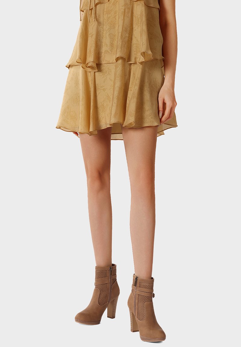 Mujer con un mini vestido de capas color marrón claro y botines de tacón alto color beige con cremalleras laterales sobre un fondo liso.
