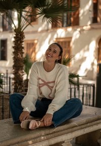 Knit ivory sweater with pink pattern, relaxed blue jeans, and pale pink ballet flats; palm tree in background, sitting on a stone bench.
