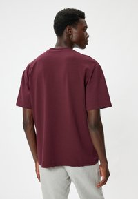 Bordeaux korteblauwe shirt met een gestructureerde stof, comfortabele pasvorm, ronde halslijn en zijsplitten, gedragen over lichtgrijze broeken.