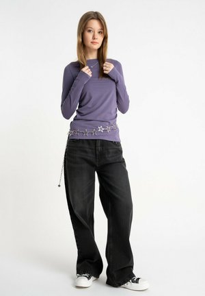 Młoda kobieta ubrana w fioletową koszulę z długim rękawem, czarne szerokie dżinsy, białe trampki oraz srebrny pasek z łańcuszka w kształcie gwiazdy, stojąca na jednolitym tle.