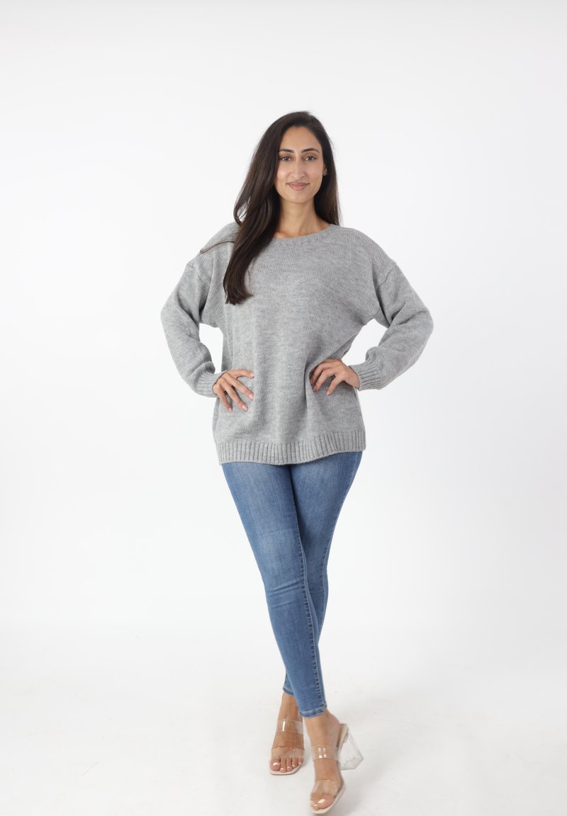 Szary, oversize sweter z prążkowanym dołem i mankietami, w połączeniu z dopasowanymi niebieskimi dżinsami i przezroczystymi sandałami na obcasie. Gładkie białe tło.