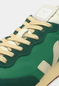 Πράσινο και εκρού αθλητικό παπούτσι με εκρού κορδόνια, λεπτομέρειες από σουέτ και λογότυπο «Veja» στη γλώσσα και το πλαϊνό πάνελ, στενή γωνιακή όψη.