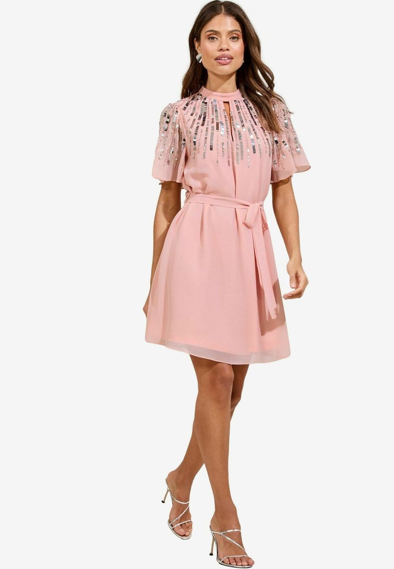 Kurzes pinkes Kleid mit kurzen Ärmeln, verziert mit Pailletten, einem taillierten Gürtel und einem knielangen A-Linien-Rock. Silberne High Heels.
