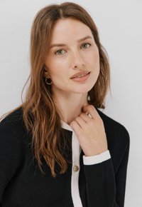 Czarny kardigan z białym kołnierzykiem i mankietami, ozdobiony złotym guzikiem. Modelka ma długie, falujące brązowe włosy i nosi złote kolczyki w kształcie kół.