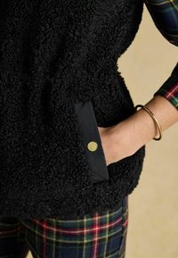 Czarna kamizelka z polaru o teksturowanym, puszystym wykończeniu oraz kieszenią z akcentem w postaci złotego zatrzasku. Noszona z spodniami w kratę.