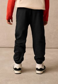 Schwarze Jogginghosen mit elastischen Bündchen und Seitentaschen, kombiniert mit weiß-schwarzen Sneakern. Das Oberteil ist hellbeige mit roten Ärmeln.