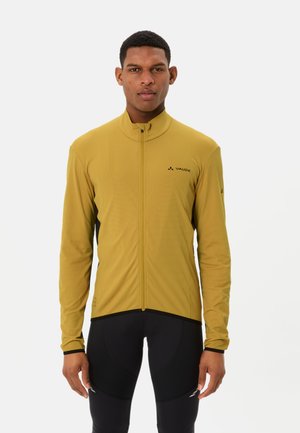 Uomo in piedi di fronte, indossa una giacca gialla senape con cerniera e pantaloni sportivi neri su uno sfondo bianco uniforme.