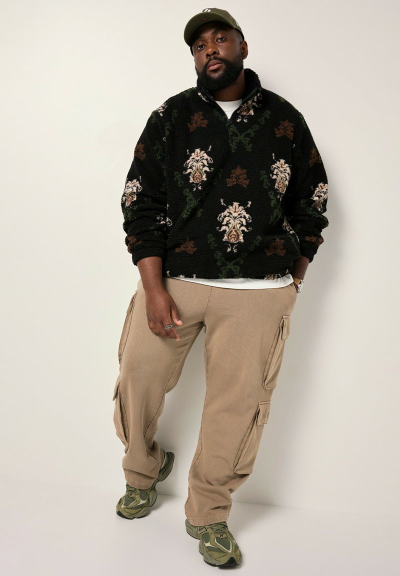 Schwarzer Fleece-Pullover mit floralem Muster in Creme, Braun und Grün, khakifarbene Cargo-Hosen mit Seitentaschen und olivgrüne Sneakers.
