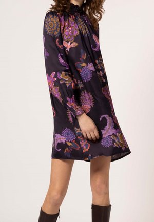 Kobieta ubrana w czarną sukienkę z długim rękawem, sięgającą do kolan, z fioletowymi, pomarańczowymi i różowymi kwiatowymi wzorami, dobraną do ciemnych kozakiów za kolano.