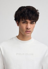 Vit sweatshirt med rund halsringning, prydd med subtil brodering av "POLO CLUB" i en ljus färg. Slät textur och vanlig passform.
