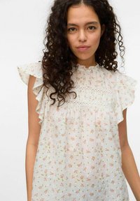 Hvid bluse i stof med små lyserøde blomsterprint, flæset korte ærmer og en delikat blondeaccent omkring brystet. Let og løstsiddende pasform.