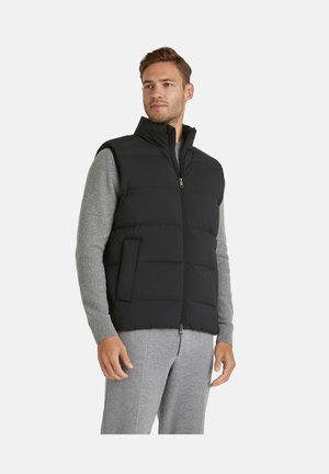 Uomo che indossa un gilet nero trapuntato senza maniche sopra un maglione grigio a maniche lunghe e pantaloni grigi, in piedi con un'espressione neutra.