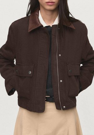 Personne portant une veste marron foncé avec grandes poches boutonnées et col en cuir, sur une chemise blanche et une jupe beige.