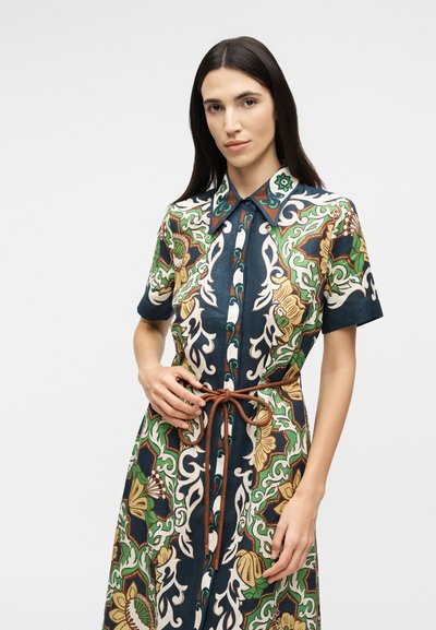 Γυναίκα με μακριά σκούρα μαλλιά, που φοράει φόρεμα με κοντά μανίκια και γιακά, με πράσινα, μπεζ και ναυτικά λουλουδένια και γεωμετρικά σχέδια, δεμένο στη μέση με καφέ κορδόνι.