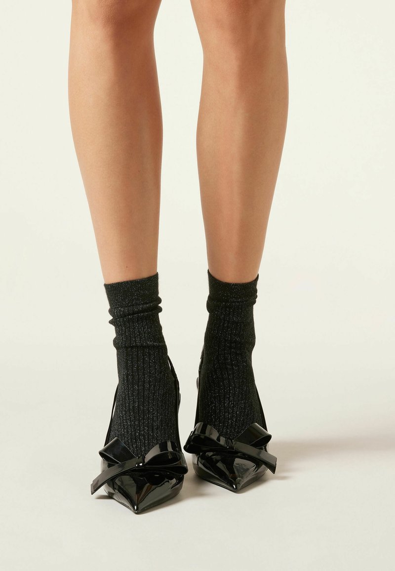 Zapatos de charol negro con punta afilada que presentan grandes lazos en cada punta. Combinados con medias negras acanaladas brillantes. Textura suave.