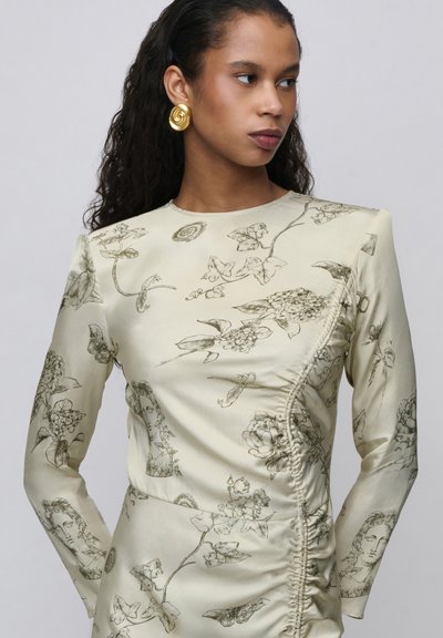 Femme aux longs cheveux bouclés portant une robe crème avec des croquis botaniques noirs et des sculptures classiques ainsi que de grandes boucles d’oreilles dorées en spirale.