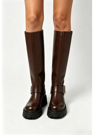 Bottes en cuir marron foncé montant jusqu'au genou avec des semelles noires épaisses et de grandes boucles, portées sur des jambes nues sur un fond uni.