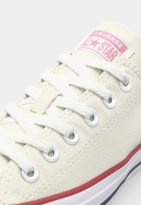 Beige canvas sneaker with white laces, silver grommets, and a pink All Star patch. Features a red and blue rubber sole.