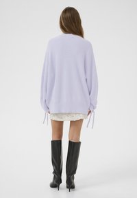 Lila, übergroßer Strickpullover mit gerippter Textur und gebundenen Ärmeln, kombiniert mit einem kremfarbenen Spitzensminirock und knielangen schwarzen stiletto Stiefeln.
