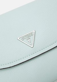 Světle modrá kabelka z faux kůže s texturovaným povrchem. Obsahuje stříbrnou trojúhelníkovou nášivku s embosovaným nápisem "GUESS USA".