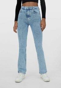 Jeans de campana en denim de algodón de color azul claro con cintura alta, cinco bolsillos y áreas deslavadas. Combinados con zapatillas blancas.