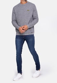 Grauer strukturierter Pullover mit langen Ärmeln, Rundhalsausschnitt, versehen mit einem kleinen gelben Emblem auf der Brust, kombiniert mit blauen Skinny Jeans und weißen Sneakers.
