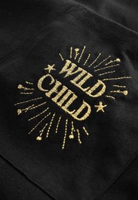 Črn material z zlatim vezene besedilo "WILD CHILD" z zvezdnimi žarki in dekorativnimi poudarki. Gladka tekstura s poliranim zaključkom.