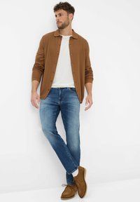Bruine overhemd met knopen over een witte T-shirt met ronde hals, gecombineerd met lichtblauwe jeans en bruine suède schoenen, tegen een witte achtergrond.