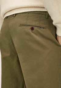 Olivegrüne Baumwollhose mit einer Gesäßtasche, die einen braunen Knopf und ein kleines "Hackett"-Logo-Label über der Tasche hat.