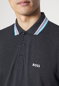 Uhlíková polo tričko s klasickým límcem, třemi černými knoflíky a bílým a tyrkysovým pruhovaným akcentem. Obsahuje vyšívané logo "BOSS".