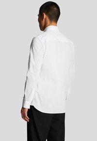 Camicia bianca a maniche lunghe con colletto classico, polsini con bottoni e una texture liscia. Design aderente che mostra la camicia da dietro.