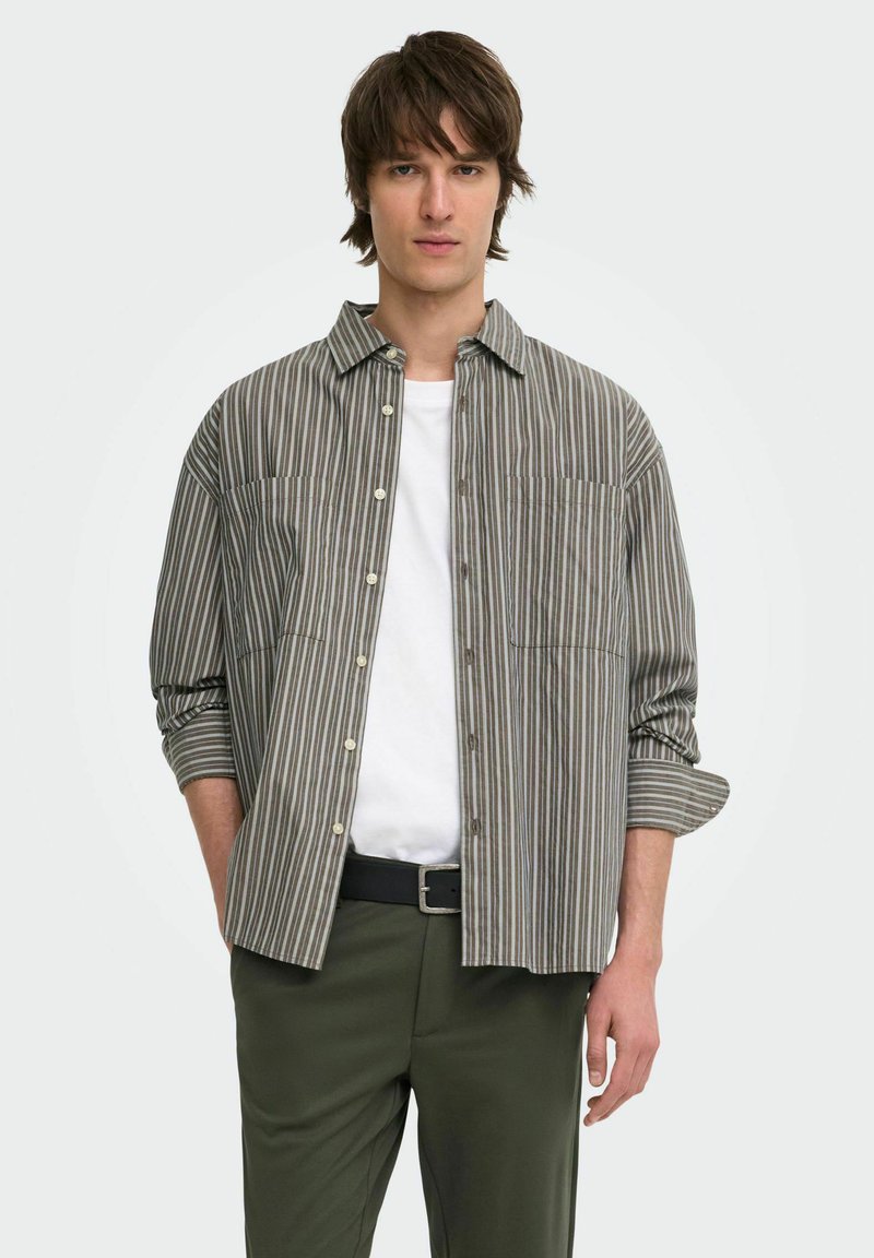 Jeune homme aux cheveux bruns portant une chemise grise rayée à boutons par-dessus un t-shirt blanc et un pantalon vert avec une ceinture noire.