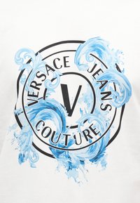 Λευκό ύφασμα με μαύρο κυκλικό λογότυπο που αναγράφει «Versace Jeans Couture» και μπλε σχέδια σταγόνων νερού που περιστρέφονται γύρω από το κείμενο και το κεντρικό γράμμα "V".