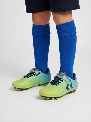 Enfant portant des crampons jaune-vert vif avec des lacets noirs et des chaussettes de football bleues remontées jusqu’aux genoux, debout sur un fond uni.