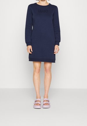 Femme portant une robe à manches longues bleu marine avec le texte "LIU JO" et des sandales roses à boucle, debout devant un fond clair uni.