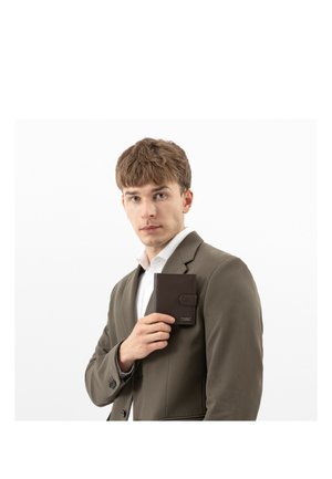 Młody mężczyzna w brązowym garniturze trzymający ciemnobrązowy portfel na wysokości klatki piersiowej na jednolitym białym tle.