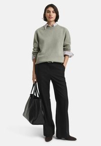Donna che indossa un maglione grigio chiaro sopra una camicia a righe, pantaloni neri a gamba larga, tiene in mano una borsa nera con texture, in piedi con una mano in tasca.