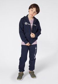Chándal navy con sudadera con cremallera y detalles de logo. Tejido cómodo, corte relajado, con puños elásticos. Combinado con zapatillas grises y verdes.