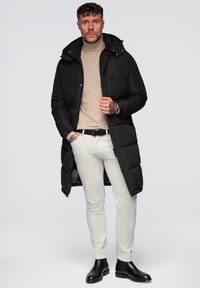 Sort polstret jakke med hætte, beige turtleneck, hvide slim-fit bukser og sorte ankelstøvler på en neutral baggrund.
