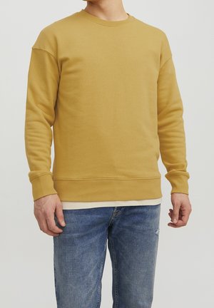 Homme portant un sweat-shirt jaune moutarde à col rond sur une chemise blanche avec un jean bleu sur fond uni.