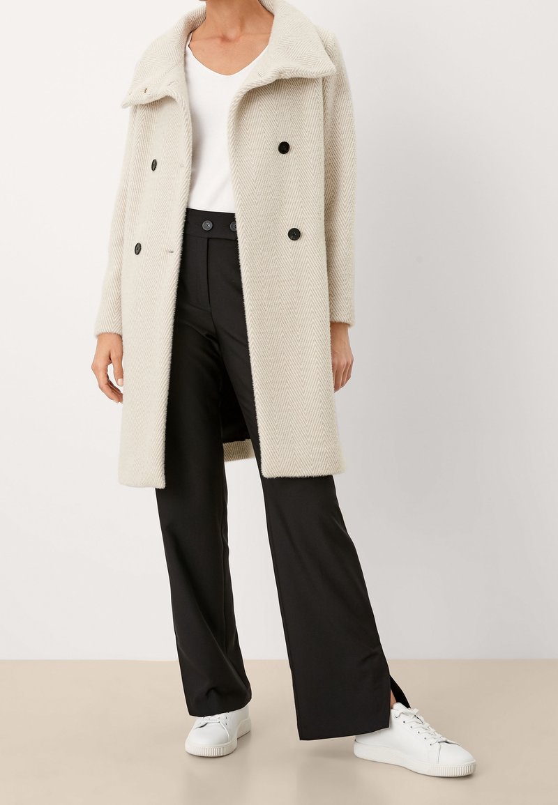 Personne portant un manteau croisé beige clair, un haut blanc, un pantalon noir taille haute avec des fentes latérales et des baskets blanches, debout.