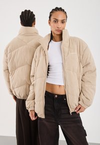Veste en peluche beige en velours côtelé avec un col haut et une fermeture éclair. Texture à côtes verticales avec un design matelassé, portée sur un haut blanc.
