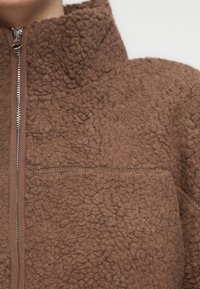 Καφέ φλις μπουφάν με υψηλό γιακά. Διαθέτει φερμουάρ σε όλο το μήκος και υφή με ανάγλυφη επιφάνεια. Απλός σχεδιασμός χωρίς εμφανή σχέδια ή στολίδια.