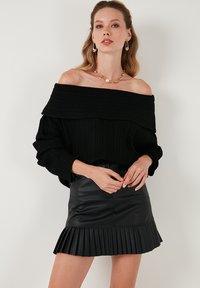 Suéter negro acanalado sin hombros con escote amplio; combinado con una minifalda negra de cuero sintético plisada. Acentos de joyería dorada.